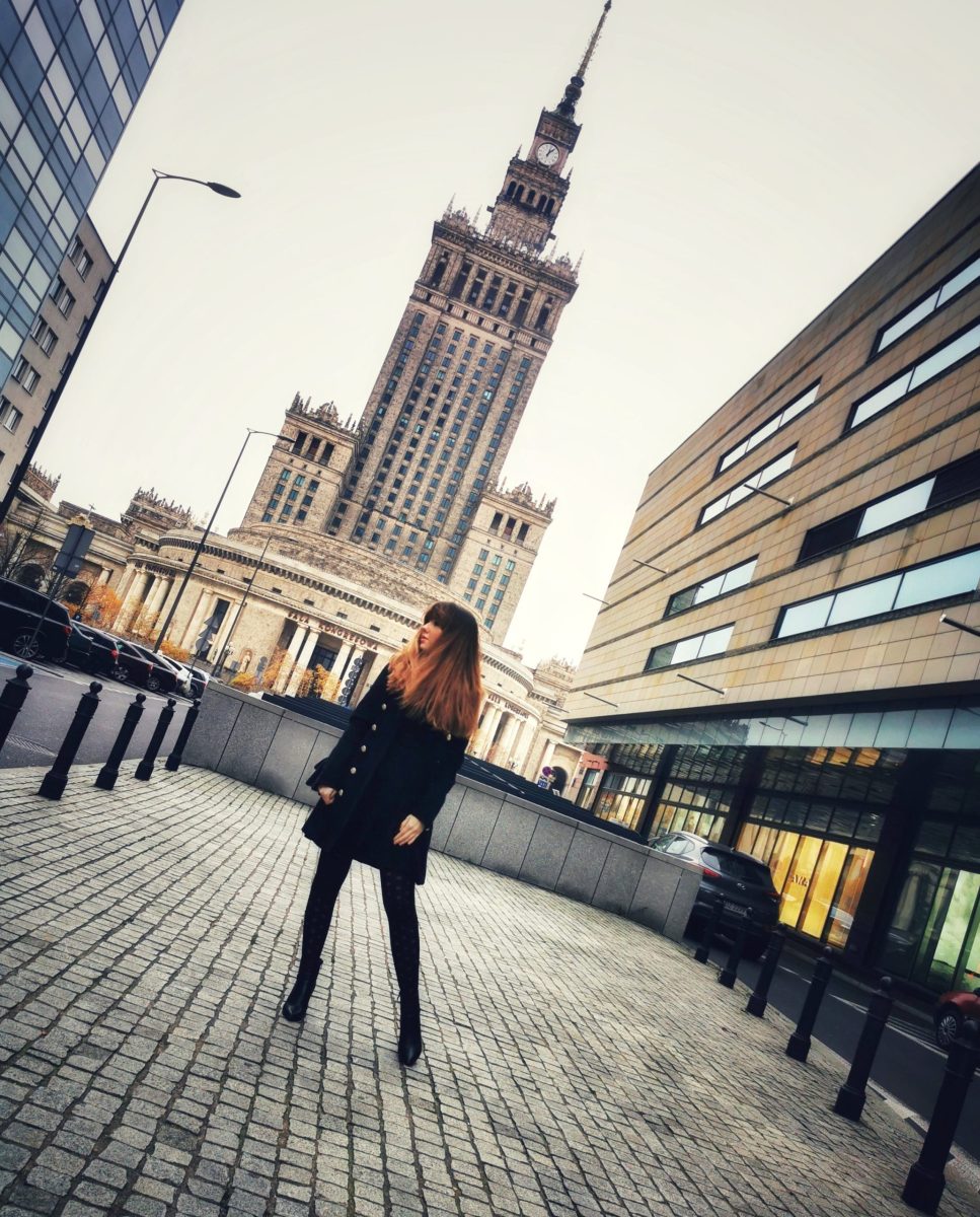 Kobieta w czarnym płaszczu zimowym na tle Pałacu Kultury i Nauki.